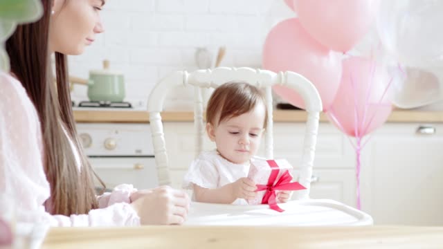 Liebenswerte-Baby-Mädchen-sitzt-im-Hochstuhl,-hält-Geschenk-für-ihren-ersten-Geburtstag-und-geben-es-zu-Mama.-Hausküche-mit-rosa-Luftballons-dekoriert