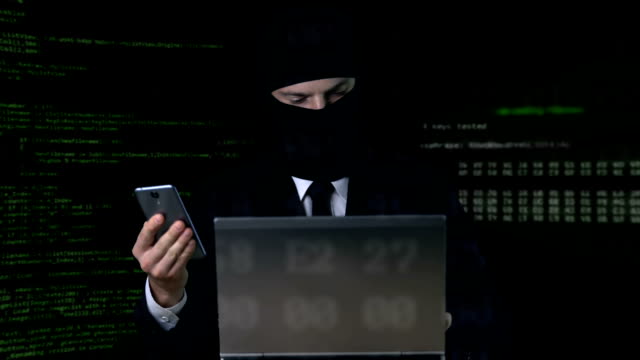 Criminal-in-suit-and-balaclava-breaking-security-code-using-laptop-and-phone