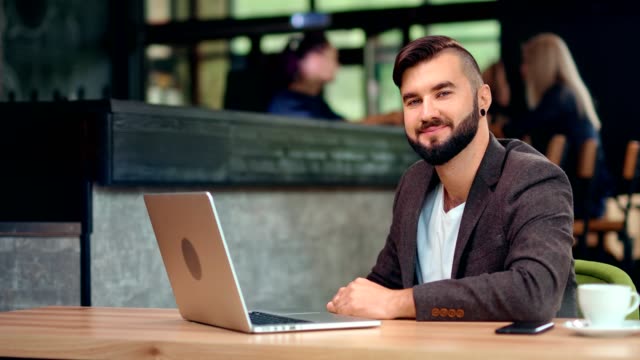Attractive-smiling-male-businessman-posing-sitting-on-table-working-using-laptop-at-cafe