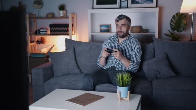 Bärtiger-Kerl-genießen-Videospiel-zu-Hause-in-der-Nacht-auf-Sofa-halten-Joystick