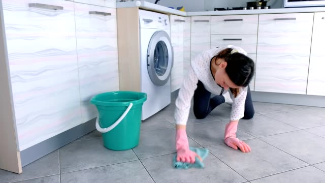 Müde-Frau-in-rosa-Gummihandschuhen-wärst-den-Küchenboden-mit-einem-Tuch.-Graue-Fliesen-auf-dem-Boden.