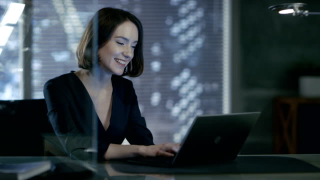 Weibliche-Führungskraft-arbeitet-auf-einem-Laptop-in-ihrem-privaten-Büro-mit-Blick-auf-die-große-Stadt.-Sie-ist-charmant-lachen.-Ihrem-Arbeitsbereich-erfolgt-in-dunklen-Obertöne.