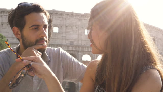Turistas-de-la-feliz-pareja-jóvenes-sentados-en-el-bar-restaurante-frente-Coliseo-en-Roma,-en-la-puesta-de-sol-charlando-y-riendo