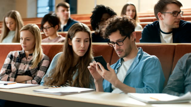 Estudiante-masculino-acciones-pantalla-de-teléfono-móvil-con-estudiante-del-compañero-durante-la-Conferencia.-Uso-de-medios-sociales-en-las-aulas-universitarias.