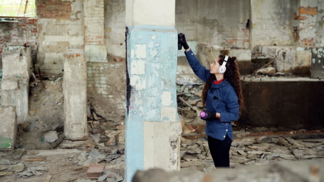Serious-pretty-girl-is-busy-painting-graffiti-on-old-dirty-pillar-in-empty-industrial-building-using-spray-paint-while-listening-to-music-with-headphones.