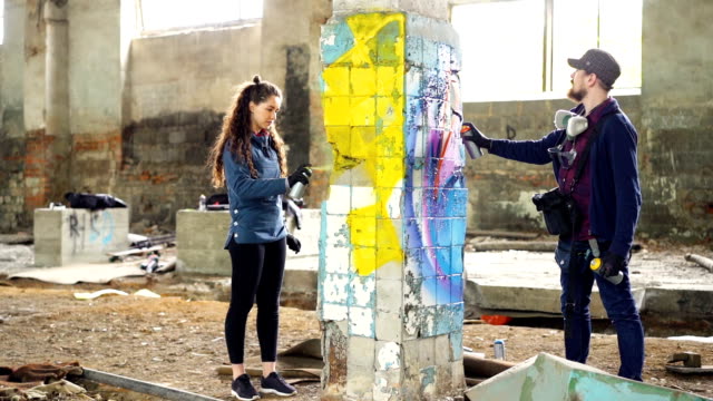 Slow-motion-of-skilled-graffiti-artists-bearded-guy-and-attractive-young-woman-working-together-in-abandoned-warehouse-decorating-damaged-column-with-abstract-image.