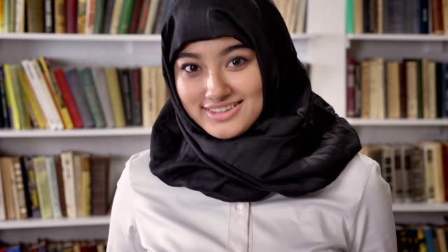 Joven-bonita-de-las-mujeres-musulmanas-en-hijab-mirando-a-cámara-y-sonriente,-de-pie-en-la-biblioteca,-estantes-con-fondo-de-libros