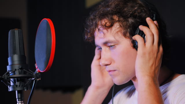 Young-handsome-singer-man-puts-on-headphones-in-the-studio.-Recording-new-melody-or-album.-Male-vocal-artist-with-curly-hair-preparing-for-working.-4k