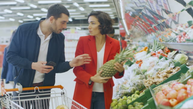 En-el-supermercado:-feliz-pareja-realiza-compras,-elegir-frutas-y-verduras-en-fresco-producen-sección.-Hombre-utiliza-Smartphone-y-empuja-el-carrito-de-compras,-mujer-coloca-productos-en-carro.