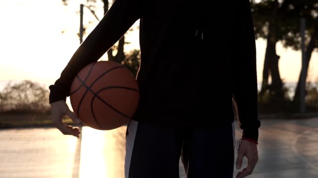 Aufnahmen-von-unten-und-oben-unten-ein-in-voller-Länge-von-einem-jungen-weiblichen-Basketballspieler-in-Hoodie-und-Shorts,-stehend-auf-einem-lokalen-Basketballfeld-mit-Ball-in-der-Hand.-Morgen-Zeit