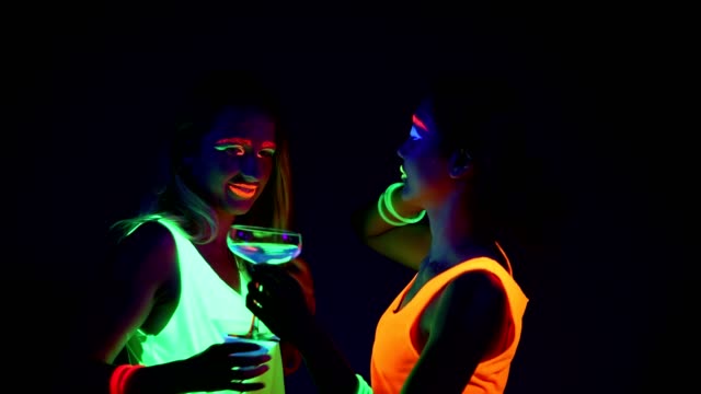 Women-with-UV-face-paint,-laser,-glowing-bracelets,-drinks,-glowing-clothing-dancing-together-in-front-of-camera,-Half-body-shot.-Caucasian-and-asian-woman.-.