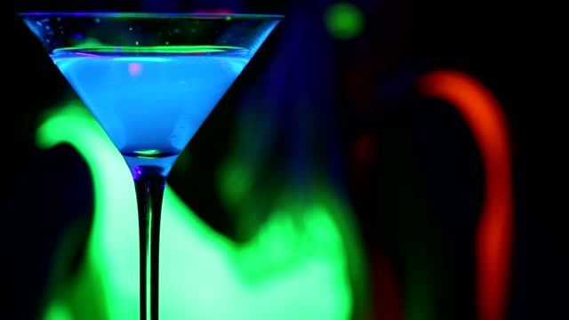Bebida-azul-que-brilla-intensamente-con-mujeres-baila-en-UV-brillo-fondo-partido-y-mujer-ven-y-coge-la-bebida.-.