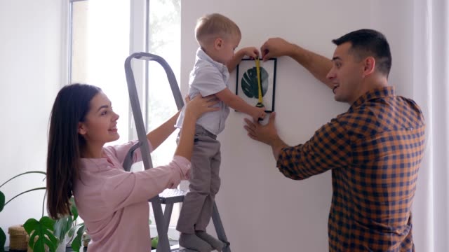 home-repair,-cute-child-with-measuring-tape-helps-to-parents-make-repairs-and-hang-picture-in-apartment