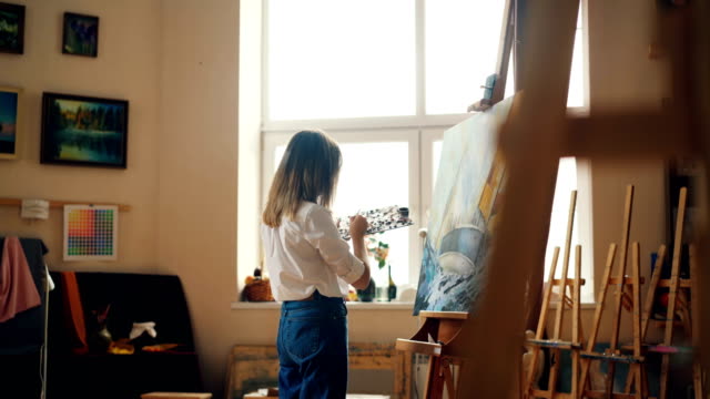 Professional-painter-young-lady-is-painting-seascape-with-acrylic-paints-depicting-marine-landscape-ship-and-sea-waves-working-alone-in-studio.-People-and-work-concept.