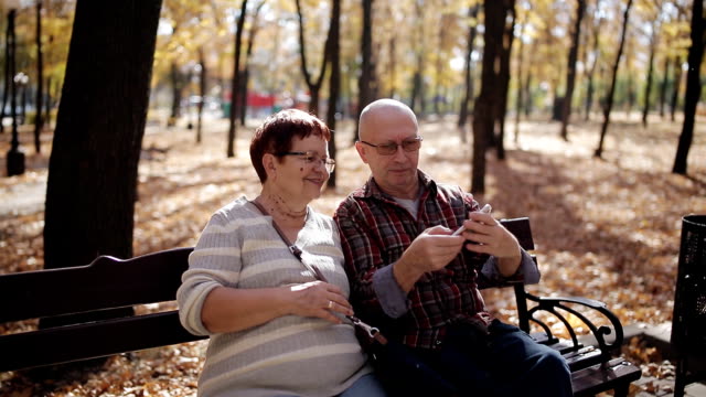 Una-pareja-de-ancianos-en-el-otoño-Parque-sentado-en-el-banquillo-tomando-autorretratos-en-un-smartphone.