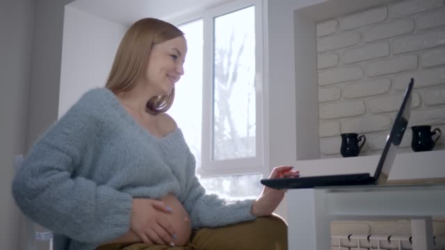 conexión-de-vídeo,-mujer-embarazada-utiliza-el-ordenador-para-chatear-con-amigos-en-Internet-y-muestra-el-vientre-desnudo-sentado-en-silla
