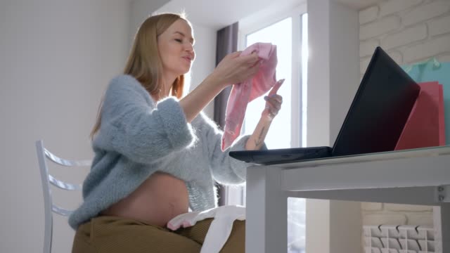 esperando-a-bebe,-mujer-embarazada-con-abdomen-desnudo-considerando-ropa-nueva-para-niño-futuro-comprado-en-Internet-sentado-frente-la-computadora