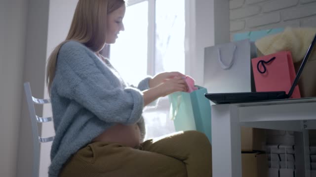 care-of-future-child,-happy-maternity-girl-with-bare-belly-examines-clothes-Internet-buying-for-an-future-baby-sitting-in-front-of-laptop