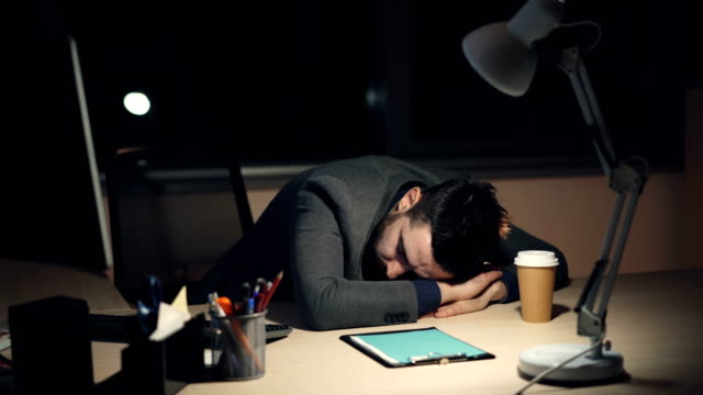 Cansado-chico-en-traje-duerme-en-escritorio-en-la-oficina-por-la-noche-después-del-ajetreado-día-de-trabajo.-Ordenador-encenderlo,-café-para-llevar-y-la-lámpara-son-visibles-en-la-tabla.