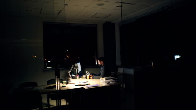 Cansado-empresario-de-traje-y-corbata-está-utilizando-computadoras-en-la-oficina-trabajando-en-la-noche-sentado-en-la-mesa-de-trabajo-en-habitación-oscura,-mirando-la-pantalla-y-escribiendo-y-tomando-café.