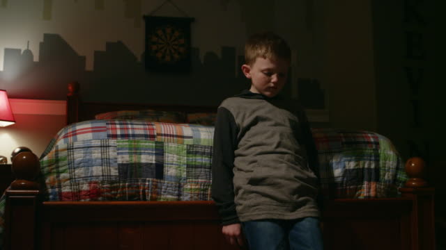 Sad-young-boy-stands-at-the-foot-of-his-bed,-crosses-his-arms