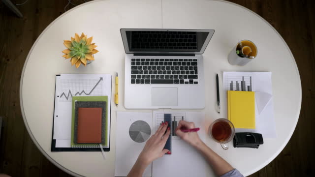 Woman-working-with-a-business-graph-and-a-laptop-that-stands-on-a-white-table-with-a-couple-tea.-Top-view.-Hands-close-up