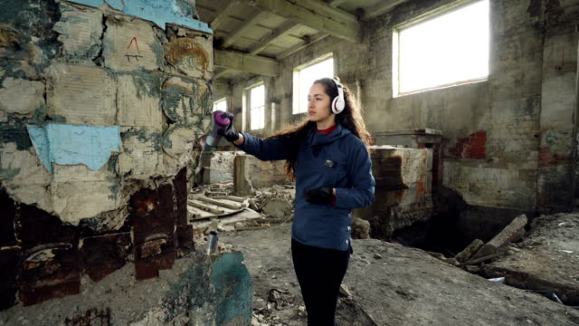 Pretty-young-woman-graffiti-artist-is-painting-with-spray-paint-inside-old-warehouse-and-listening-to-music-through-wireless-headphones.-Youth-subculture-concept.