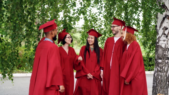 Joyful-graduating-students-multiracial-group-are-putting-hands-together-then-clapping-hands-celebrating-graduation,-friends-are-standing-outdoors-wearing-gowns-and-mortar-boards.