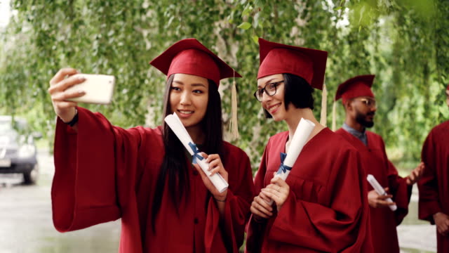 Pretty-girls-graduating-students-are-taking-selfie-with-diploma-scrolls-using-smartphone,-women-are-posing-and-smiling,-their-fellow-students-are-celebrating-in-background.