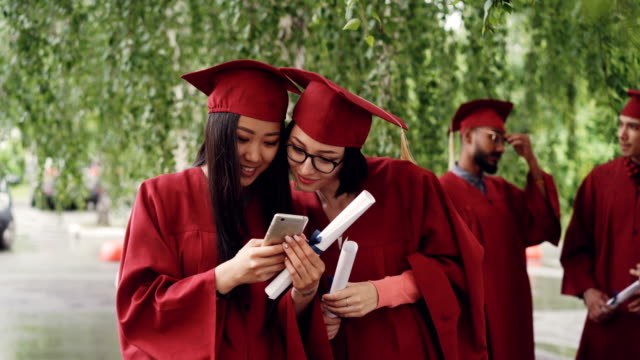 Female-graduates-are-using-smartphone-looking-at-screen-talking-and-laughing-standing-outdoors-holding-diplomas,-girls-are-wearing-formal-gowns-and-hats.
