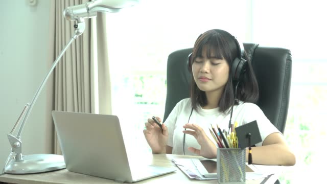 Student-Online-Studie-Lernkonzept:-erlauben-schöne-Asiatin-sitzen-Lächeln-enge-Augen-scheinen-so-gerne-hören-Musik-über-Kopfhörer-mit-ihrem-Laptop-im-Hause,-selektive-Bleistifte-und-Kugelschreiber-Box