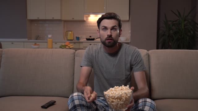 Guy-sits-on-sofa-and-watches-TV.-He-eats-popcorn.-Guy-hs-it-in-glass-bowl.-He-is-concentrated-on-watching.-He-looks-straight-forward