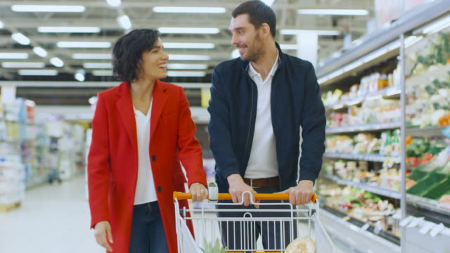 En-el-supermercado:-joven-pareja-camina-a-través-de-la-sección-de-productos-frescos-de-la-tienda-feliz,-hablar,-hombre-mujer-abraza-amorosamente.