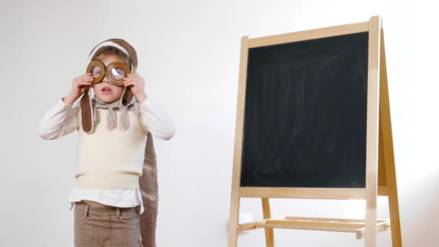 A-little-girl-dressed-as-an-airman-or-a-pilot,-indicates-with-her-hand-the-blackboard-behind-her-as-a-flight-insign-to-learn-to-use-both-aircraft-and-imagination.