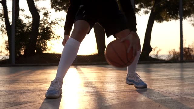 Close-up-footage-of-a-female-athlete-legs-in-white-golf-socks-and-sneakers.-Female-baasketball-player-bouncing-ball-from-hand-to-hand.,-practicing-cross-legs-exercise.-Sun-shines-on-the-background