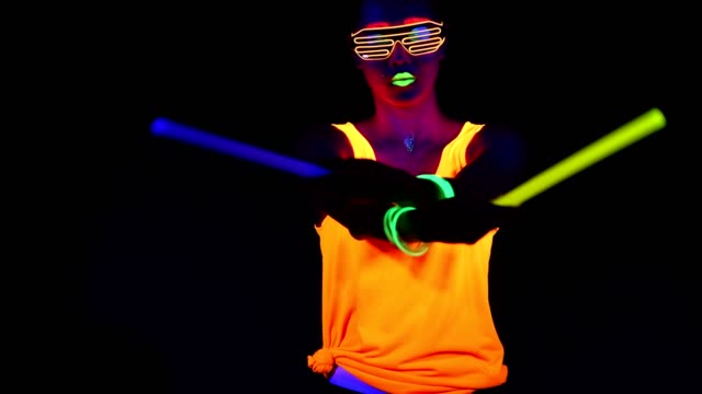 Woman-with-UV-face-paint,-glowing-clothing,-glowing-glasses,-bracelet-dancing-in-front-of-camera-holding-chemical-light,-half-body-shot.-Asian-woman.-.