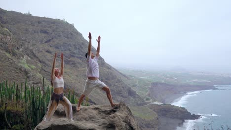 Ein-Mann-und-eine-Frau-stehen-am-Rand-einer-Klippe-mit-Blick-auf-den-Ozean-erheben-ihre-Hände-oben-und-atmen-Sie-die-frische-Seeluft-während-yoga