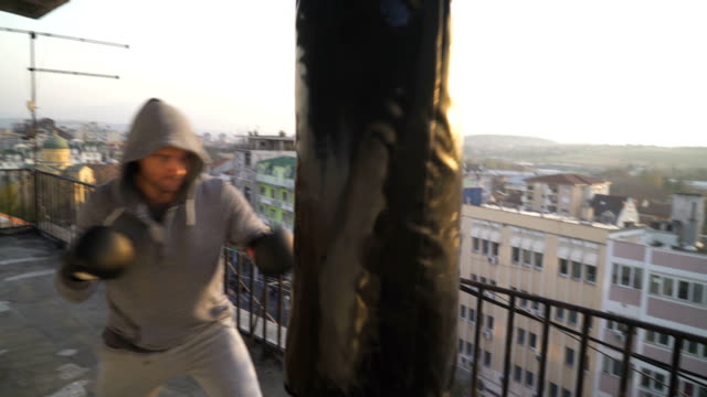 Man-with-hood-on-head-punches-bag,-boxing-on-terrace,-city-in-the-background,-sportsman-practicing,-power-training,-strong-guy-hard-exercising,-strength-exercises,-workout,-handheld,-sunny-day.--P