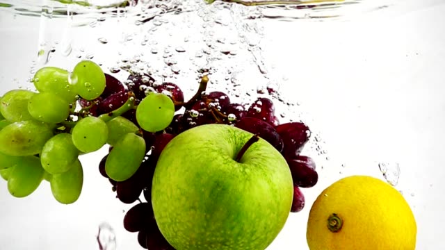 The-lemon,-apple-and-grapes-falling-in-water-with-bubbles.-Video-in-slow-motion.-Fruits-on-isolated-a-white-background.