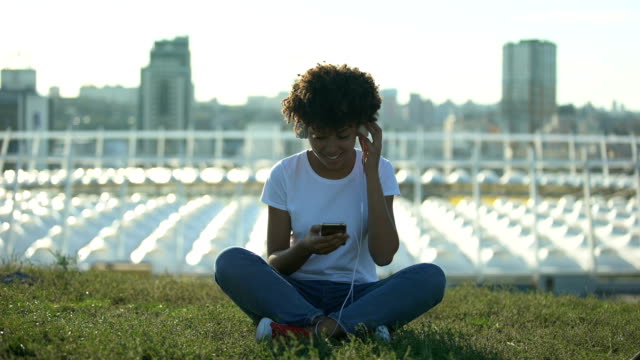 Young-afro-american-woman-listening-to-music-in-headset,-sitting-on-lawn,-relax