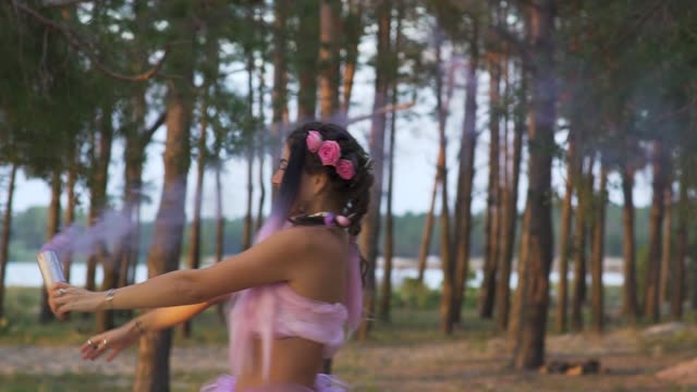 Beautiful-girl-with-bright-makeup-in-a-pink-dress-dancing-with-smoke-bombs-on-the-background-of-pine-trees.-The-dance-of-a-sensual-woman-with-a-flower-hairstyle.-Slow-motion.