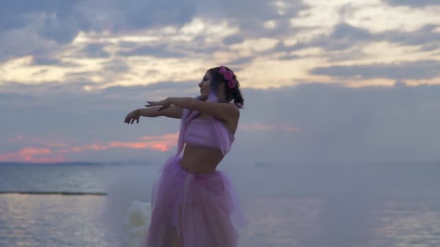 Cute-girl-with-bright-makeup-in-pink-dress-dancing-in-the-viscous-mist-from-smoke-bombs-outdoors.-The-dance-of-a-sensual-woman-with-a-flower-hairstyle.-Slow-motion.