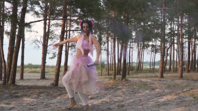 Charming-lady-with-bright-makeup-in-a-pink-dress-dancing-with-smoke-bombs-on-the-background-of-pine-trees.-The-dance-of-a-sensual-woman-with-a-flower-hairstyle.-Slow-motion.
