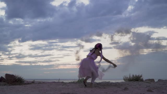 Sinnliche-junge-Frau-mit-funkelnden-Make-up-in-einem-rosa-Kleid-tanzen-mit-Rauchbomben-am-Ufer-des-Flusses.-Der-Tanz-ein-zart-süßes-Mädchen-mit-einer-Blume-Frisur.-Slow-Motion.