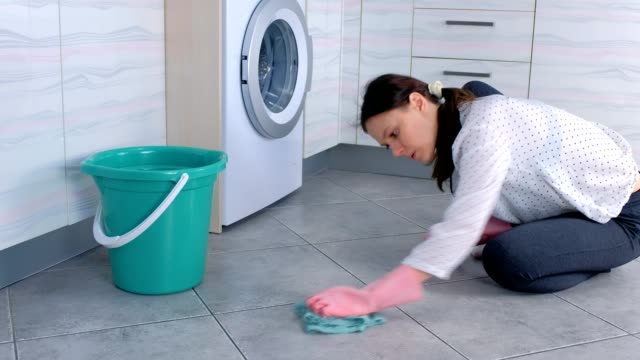 Tired-woman-in-pink-rubber-gloves-washes-and-rubs-hard-the-stain-on-kitchen-floor-with-a-cloth.-Gray-tiles-on-the-floor.