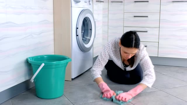 Tired-woman-in-pink-rubber-gloves-washes-kitchen-floor-with-a-cloth-and-looks-at-camera.-Gray-tiles-on-the-floor.