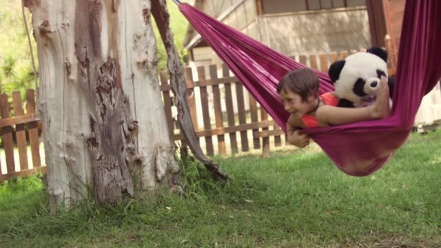 Young-happy-little-girl-in-summer-camp-lying-in-colorful-hammocks-relaxing-playing-outside-with-plushes-panda-dolls-in-green-grass-field-kids-children-slow-motion