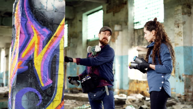 Attractive-bearded-guy-graffiti-artist-is-talking-to-his-serious-student-pretty-young-woman-and-teaching-her-how-to-use-aerosol-paint-decorating-old-building.
