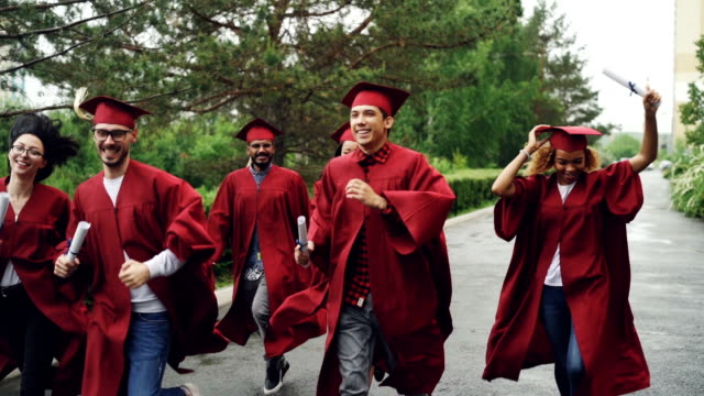 Slow-motion-of-cheerful-grads-running-together-under-rain-waving-diplomas-and-laughing-wearing-red-gowns-and-mortar-boards.-Small-rain-is-visible.
