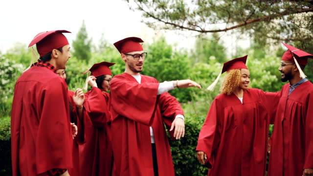 Excited-young-men-and-women-grads-are-celebrating-graduation-day-laughing,-doing-high-five,-hugging-and-dancing-outside-on-campus.-People-are-wearing-traditional-garments.